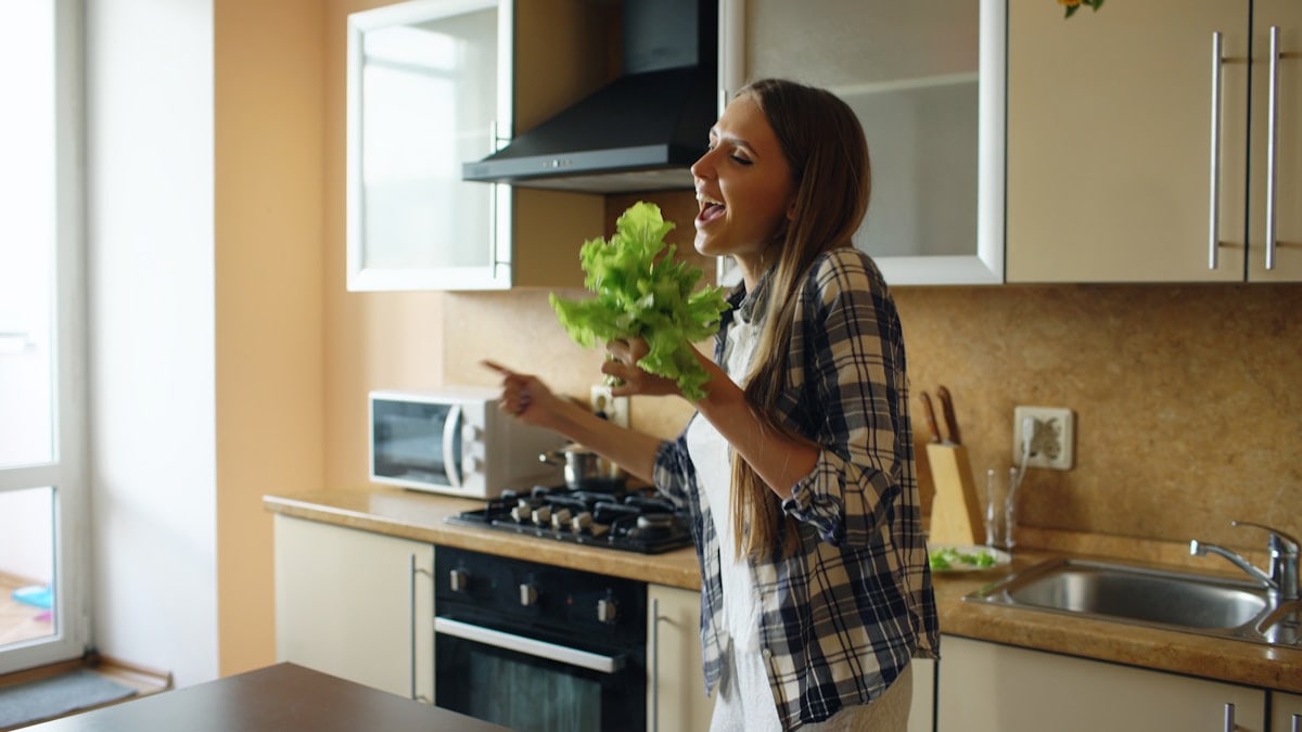 Cheerful young funny woman dancing and singing with lettuce microphone while cooking breakfast in the kitchen early morning at home